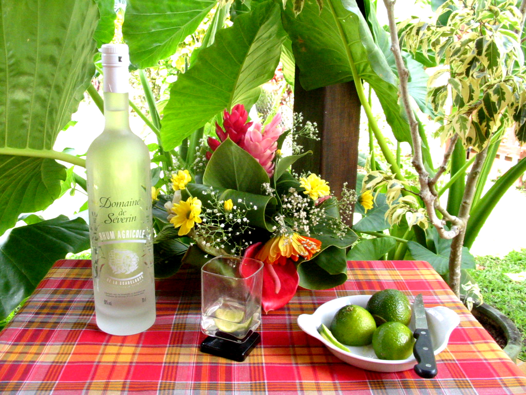 Une bouteille de rhum agricole blanc, des citrons verts et un verre de ti-punch sur une nappe madras